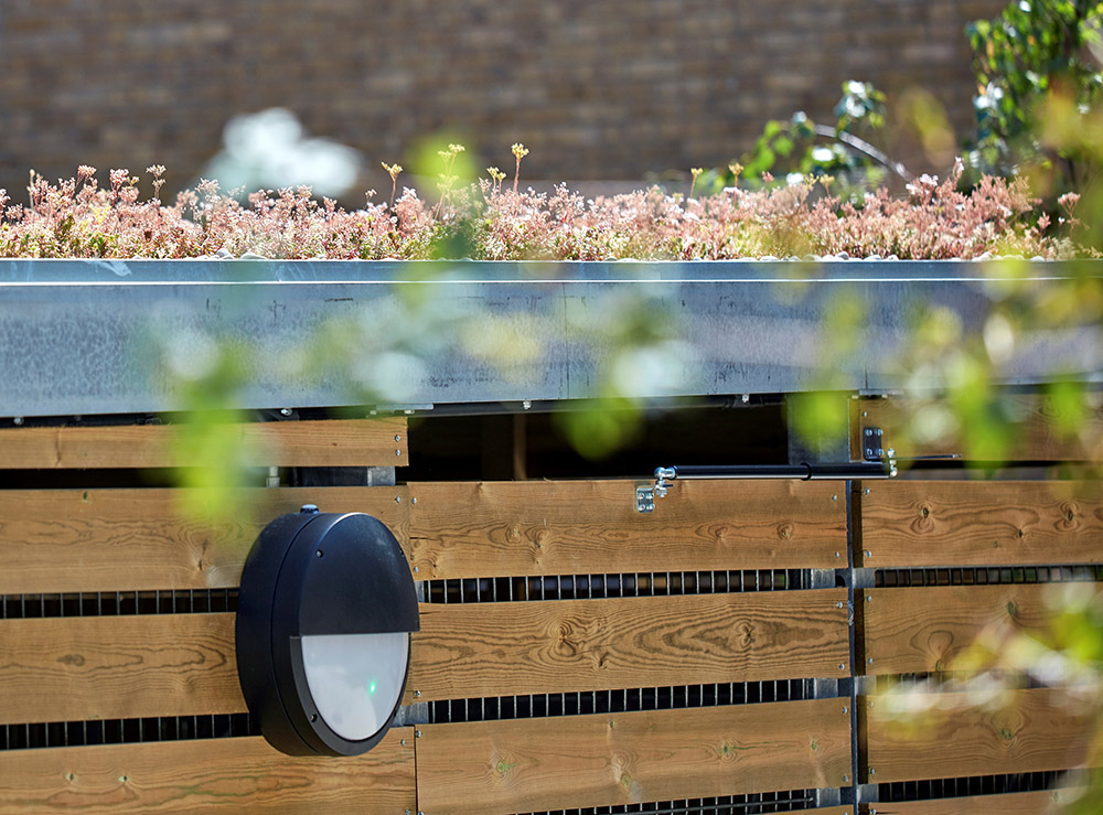 Wooden bicycle shelter with flowers on roof and light attached to side. On door is a self-closer.