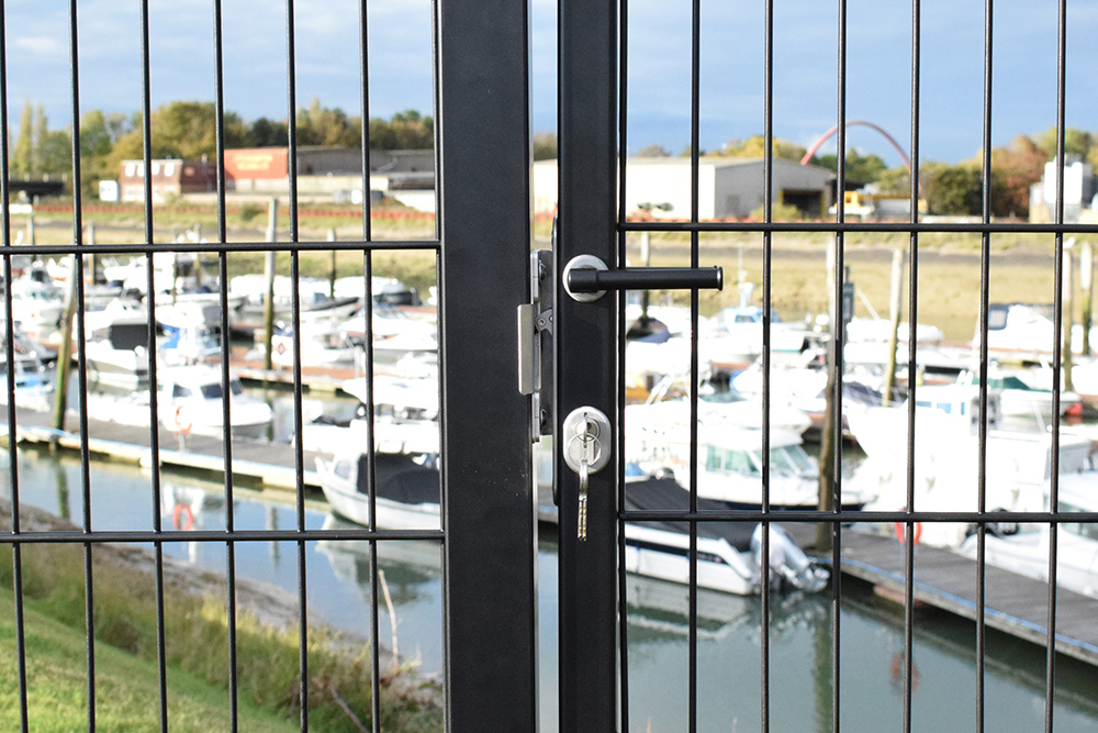 ML4 mortice hook lock installed in black metal mesh gate frame. Gate is standing in front of a marina with lots of boats and a boardwalk.