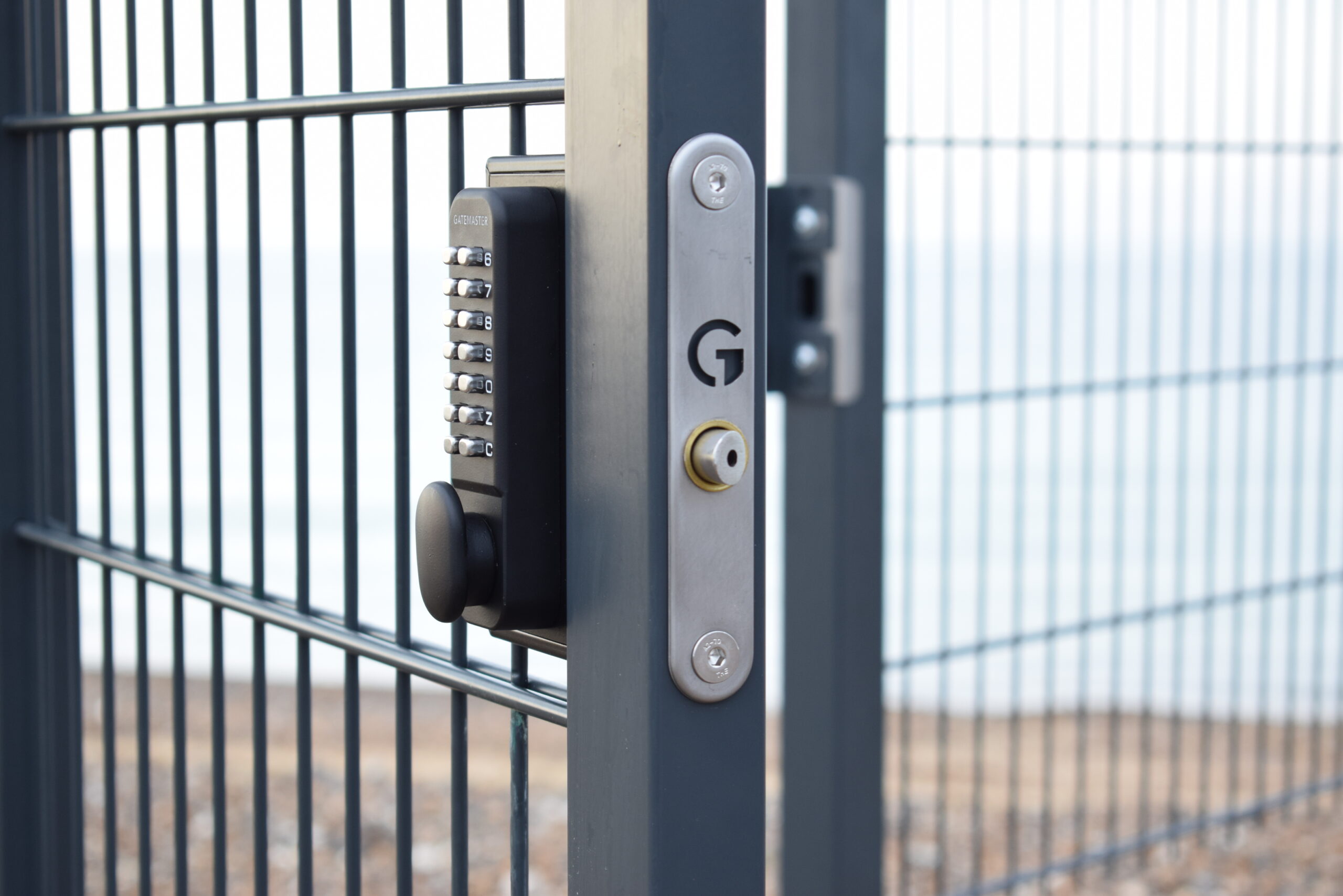 Serrure de portail à combinaison sans clé installée sur un portail en acier thermolaqué en gris. La porte est sur un hêtre avec de l'eau floutée en arrière-plan.