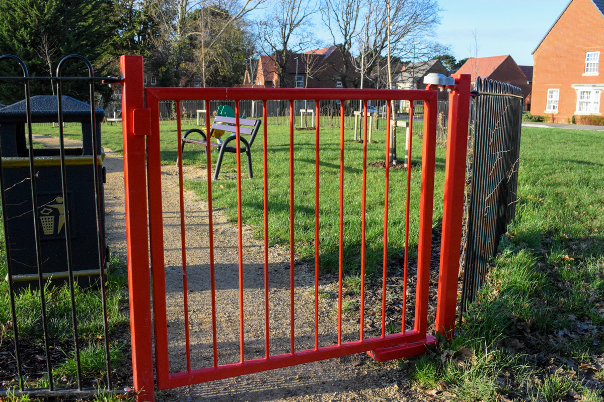Red metal gate on public park. Gate has gate closing mechanism installed and is in front of a bin and benches,