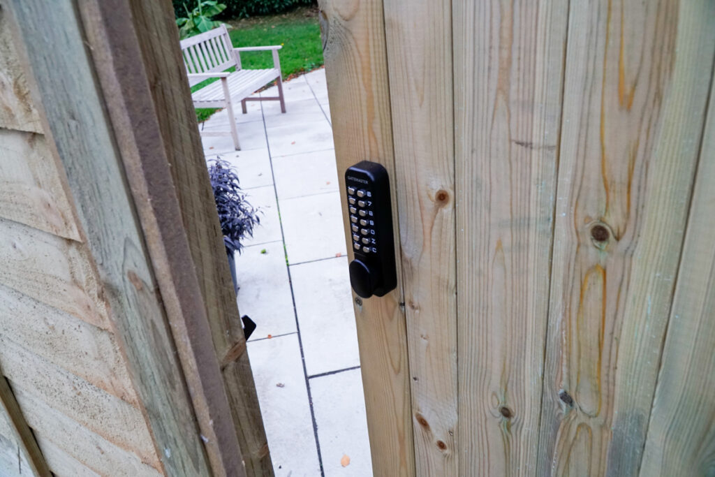 Open gate with digital lock for wooden gate showing. Through the opening, is a bench on paved path and grass.