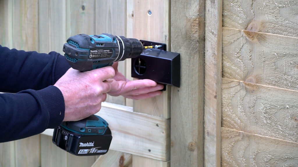 Personne avec une perceuse électrique montant un verrou à clé et un cache sur un portail de jardin en bois