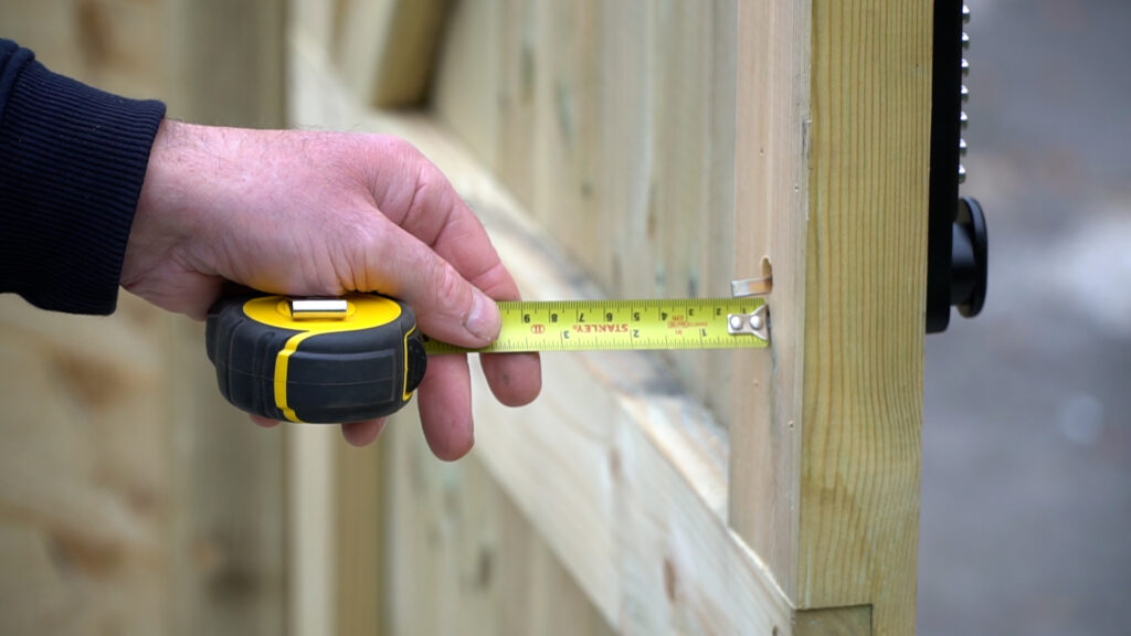 Personne mesurant une broche dépassant d'un portail en bois avec un ruban à mesurer jaune